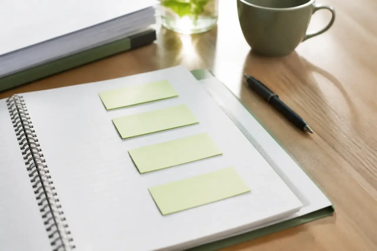 Dossier de transmission posé sur une table, post-it vierges alignés sur une page de carnet, stylo et tasse en arrière-plan