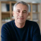 Portrait de Lucien Mérian, cheveux gris courts, pull bleu nuit, sourire discret, fond de bibliothèque lumineuse et floue