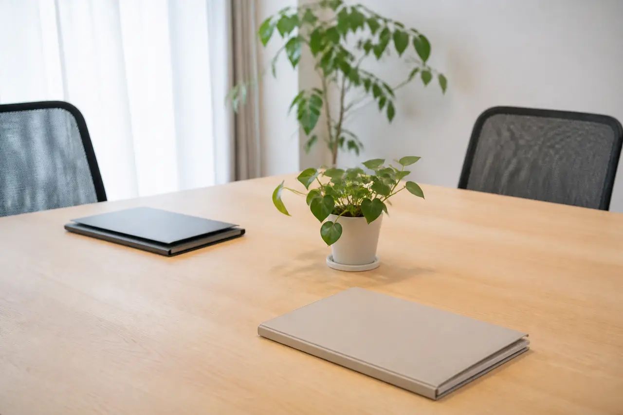 Une table de réunion en bois clair avec deux chaises vides, des dossiers fermés et une plante verte